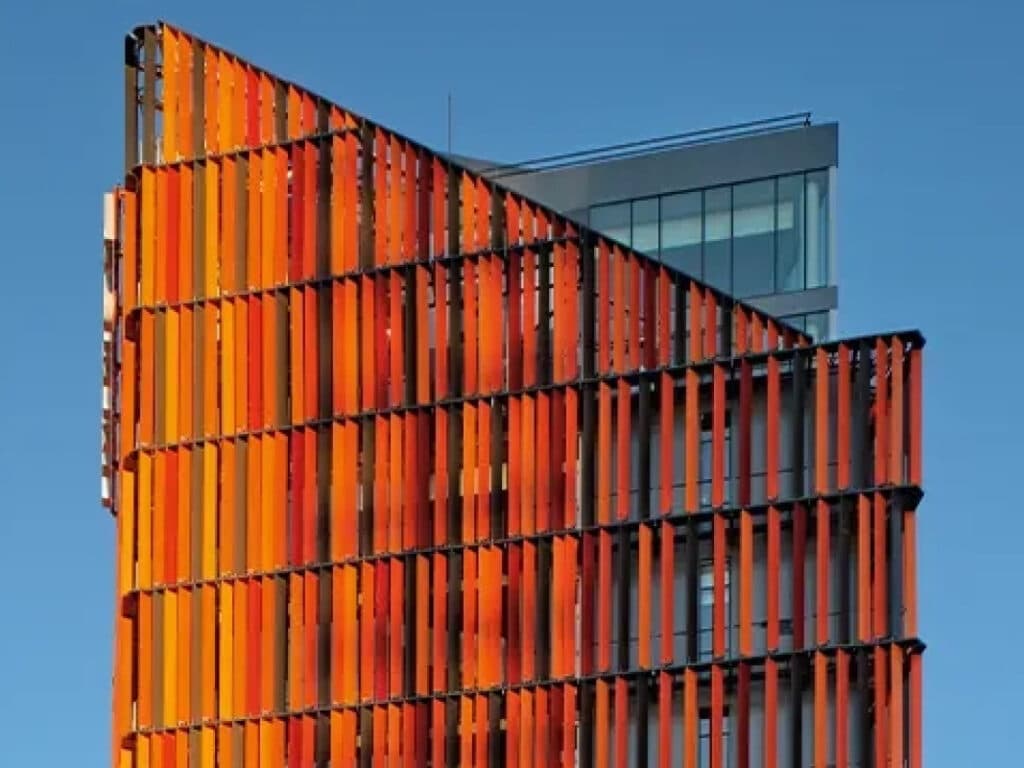 Futuristisches Gebäude mit oranger, lamellenartiger Fassade und blauen Glasfenstern vor blauem Himmel.