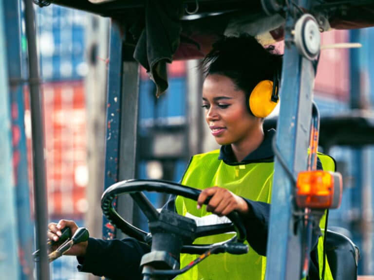 Frau in Schutzweste und Gehörschutz bedient Gabelstapler vor bunten Schiffscontainern im Hafen